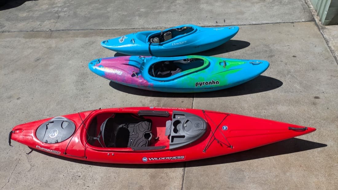 Three plastic kayaks lined up on concrete, including whitewater and touring hulls
