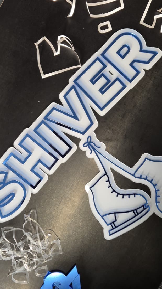Layered blue and white acrylic lettering spelling Shiver with an ice-skate motif