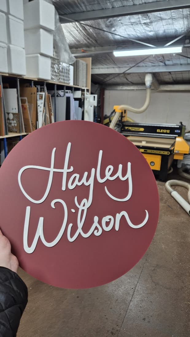 Round routed acrylic sign with white script lettering reading Hayley Wilson, held in the workshop